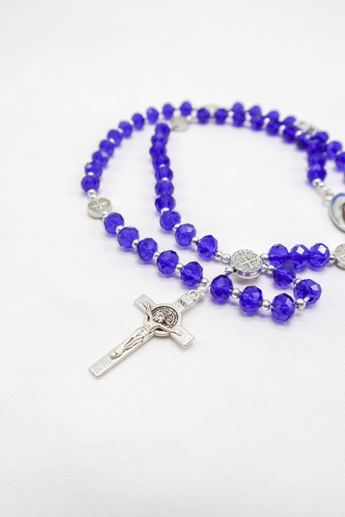 Placeholder photograph of blue rosary beads and a silver crucifix on a white background. Photo by Rasha Saadeh on Pexels; not an actual Blessed By Mary product.